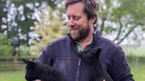 Tom Martin is stood outside in a field holding two black lambs in each arm. He wears a blue zip up fleece with a green shirt underneath and had dark brown hair and a matching colour beard. He is looking at the lambs and smiling. 