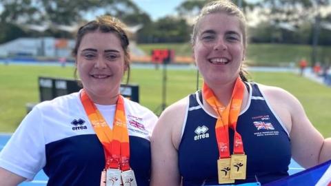 Two women wearing medals hold a British flag in front of an athletics track