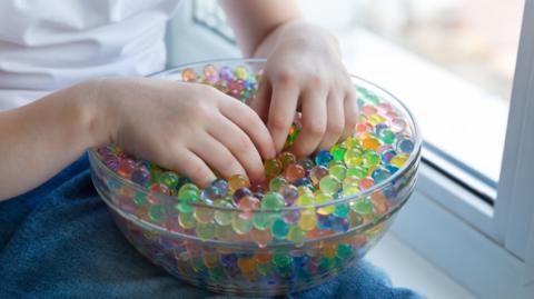 Child playing with water beads. The beads are of different colours and in a clear bowl. The hands of the child can been seen.