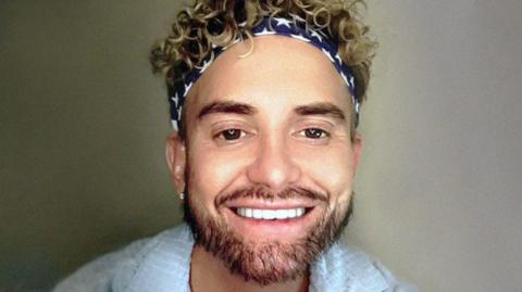 A smiling selfie of Kevin Sutherland. He is wearing a light blue shirt which is open at the neck. He has dark/ blonde curly hair and is wearing a black and white bandana with stars on it. 