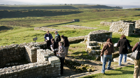 A group of people wonder through the walled remains of Housesteads Roman Fort in Northumberland 