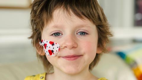 Annabel, a young girl with blue eyes, short dark hair, and a tube coming from her nose concealed by a plaster with a ladybird pattern on it.
