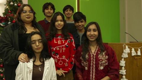 Four teenage girls and three teenage boys stand in a church with a green wall and a Christmas tree.