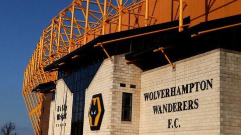 A view of the Molineux Stadium in Wolverhampton with brick walls and orange bars creating a structure on the top, with a sign on the wall saying Wolverhampton Wanderers FC and the club logo.