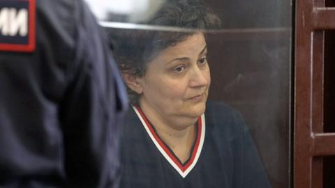 Anna sitting behind the glass screen of the court cage with a wooden frame. She is on the right side of the shot, looking towards the right with a serious expression on her face. She has short dark hair, wearing a navy v neck jumper with red trim. On the left-hand is the back of a security guard with Russian writing on his navy uniform.