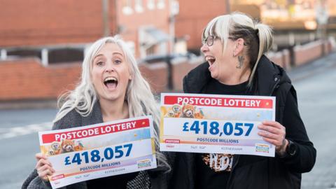 Two women standing on a street, each holding a cheque worth £418,047. On the left is Lauren Tomlinson, aged 33, who has long blonde hair and is smiling with her mouth wide. Her mum, Andrea, is on the right looking at her, smiling with her mouth open too. She has glasses and hair with blonde on top and brown underneath.