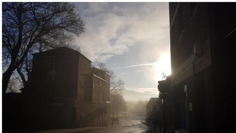 Low cloud and mist surround a street with the sun breaking through in the distance. 