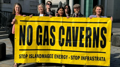 Campaigners standing outside Belfast Laganside Court complex in June 2024 holding a large yellow and black banner reading "No gas caverns"
