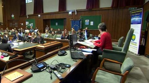 Children sit in wood panelled council chambers during a model climate conference, in their school uniforms. An older woman with short grey hair, who is wearing a long sleeved red top, sits at the desk at the front. 