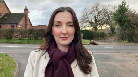 A woman with long dark hair past her shoulders wears a light white coat with a purple scarf knotted around her neck. She stands on a road leading to a main road and behind the road is a red brick building next to trees and a muddy path