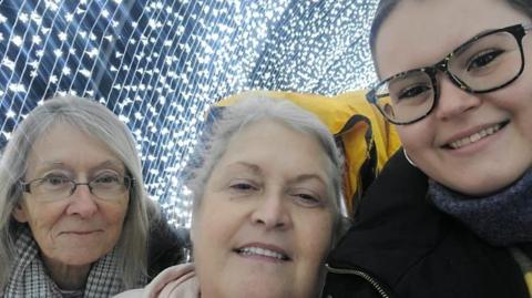 Jordan with her mum and aunt at a night-time lights show. The three women are side by side and smiling at the camera. Jordan has square framed glasses, a stripy scarf, black coat, earrings and brown hair which has been tied up. Her mum has grey hair and wears a pink jumper. Her aunty has shoulder length grey hair and a checkered print grey scarf. 