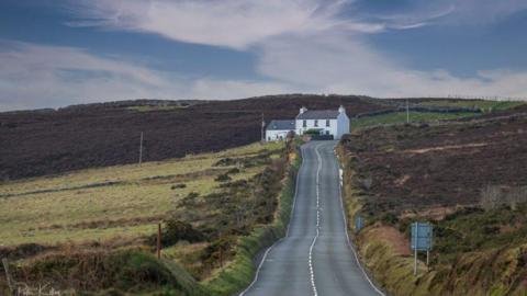 The road leading from Creg ny Baa with a grass embankment on the right, and green hills on the left. a white cottage is straight ahead on the left side of the road.