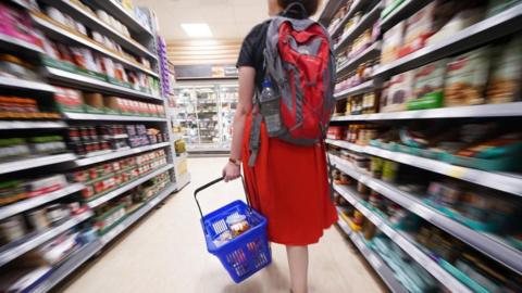 Woman in a red dress walks through aisle of supermarket with blue basket carrying food products.