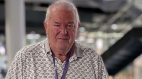 Steve Mitchell, has silver short hair, and stands centre infront of the glass rail on the first floor inside BBC central square building in Cardiff. He wears a white and grey polo shirt and has a purple visitor lanyard around his neck.