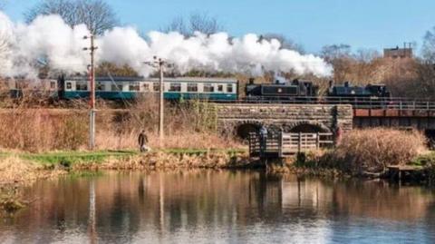 A green heritage train passing over a viaduct above a river on a sunny day with steam billowing overhead as man walks his dog along a river bank under the structure.