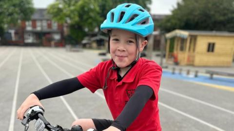 Harvey Goodman, nine, smiles at the camera while sitting on a black pushbike. He is wearing a blue bike helmet and a red polo-neck school uniform t-shirt over a long-sleeved black thermal top