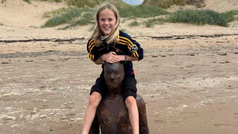 A boy with long blonde hair sits on the shoulders of one of the iron men statues on Crosby beach and smiles at the camera.