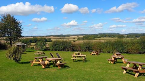 A lovely sunny day with picnic benches scattered on a grassy landscape looking out on to fields. The fields are lined with trees and hedges