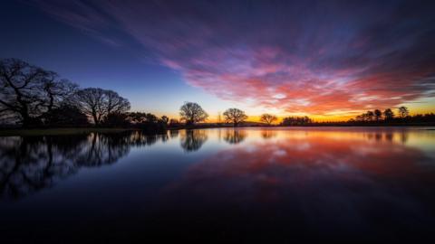 There is a large expanse of water with trees lining its distant shore. The trees are silhouetted against the sky, which is a dark blue colour but also bright orange on the horizon and peppered with pink clouds.