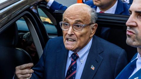 Rudy Giuliani gets out of a car at the New York Federal Courthouse on 7 November. He wears glasses, a navy suit jacket, light blue shirt and a navy and red tie patterned with stars.