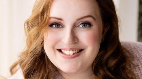 Jen has long, wavy reddish-brown hair. She is wearing a pale pink knitted cardigan and is smiling at the camera.