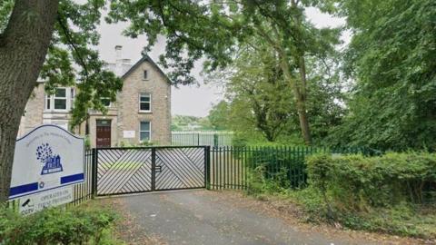 The gates of the Meadows School. It is a large three-storey grey brick house with a brown door. It is surrounded by trees and shrubs.