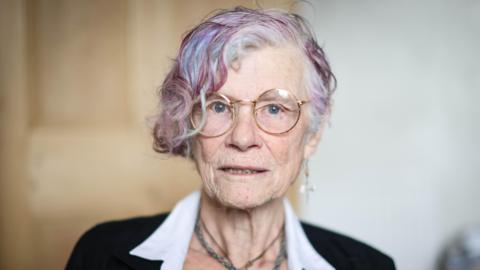 Head and shoulders image of Nikki MacLeod, who was a victim of an online romance scam. She is 77, has thin-rimmed glasses, short grey hair with purple streaks and is looking straight at the camera. She has a dangly cross earring in her left ear and is wearing a beaded necklace.