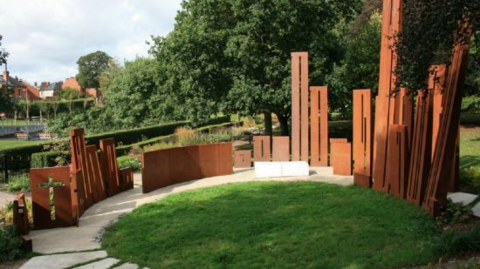 The battle memorial has been placed in a circle around the park with brown planks pointing up to the sky. In the background there are green trees and grass.