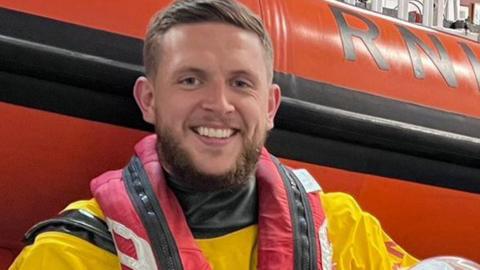 Connor Wray in his yellow lifeboat volunteer uniform and red lifebelt, holding a helmt and standing in front of an elevated orange RNLI boat in its station depot