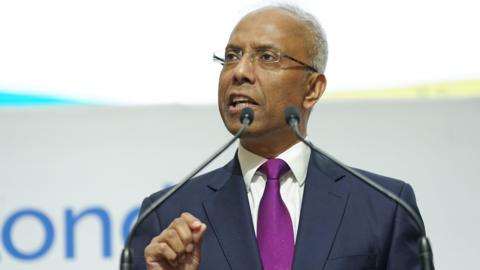 Lutfur Rahman standing on front of some microphones. He is wearing glasses, a dark blue suit with a white shirt and a purple tie. 