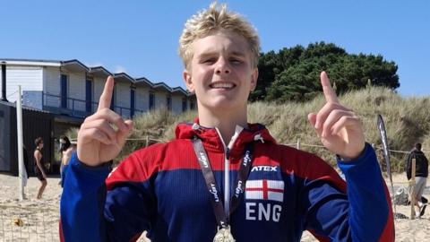 Lucas standing on a beach in a blue and red tracksuit top with a St George flag on the chest. He is smiling and pointing upwards with both hands and has a gold medal around his neck