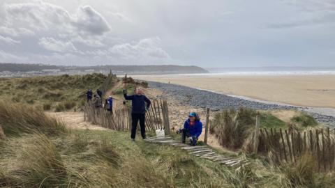 Northam Burrows rangers, two people facing the camera and five in the background, putting up a fence at the dunes at Northam Burrows. The beach stretches out behind them