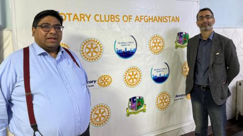 Dr Michael Fernando in a blue shirt and red braces, standing in front of a Rotary Clubs of Afghanistan banner, with another man standing to the right, wearing a grey blazer and jeans