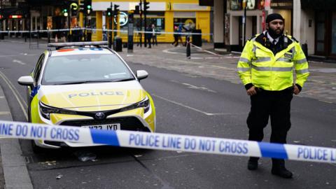 A police officer stands behind police tape near Shaftesbury avenue - a police car is also visible to the left of the image 