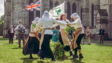 People in historical dress dancing in a circle with a village flag and oak branches