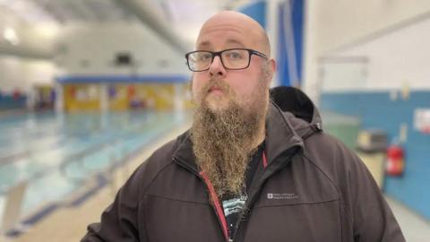 Juan McGuinness, who is bald, wears glasses and has a long beard, standing beside Ramsey Swimming Pool.