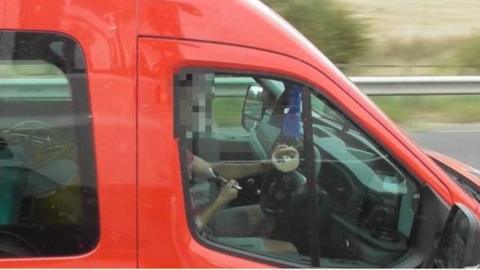 The driver of a red vehicle is seen eating from a circular bowl while holding one hand on the steering wheel.