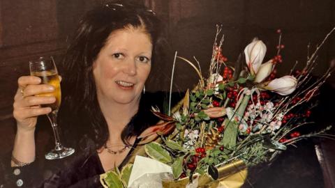 Shona Campbell, a woman with dark hair, smiling as she holds a glass of champagne and a large bouquet of flowers.