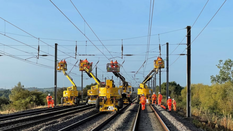 Large yellow equipment on the railway lines providing elevated platforms for workers, dressed in orange, to reach overhead lines.