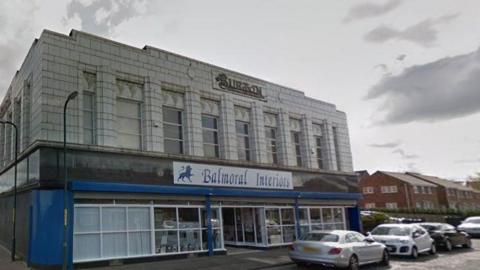 The Burton building. It is a two-storey white stone building in an Art Deco style, with a blue shop front on its ground floor. There is a Balmoral Interiors sign above the entrance and the word Burton appears in an ornate typeface above the central first floor windows. Several cars are parked on the street.
