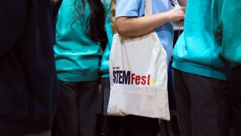 A white canvas bag with the word STEMFeston it over a school girl's shoulder. She is wearing a blue school uniform and has other students around her.