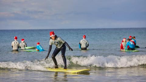 A woman, wearing a Santa hat, is surfing. The surfer is part of the Wave Project and is standing on a yellow surfboard. She is wearing a wetsuit. Other surfers in Santa hats are waist deep in the water behind.