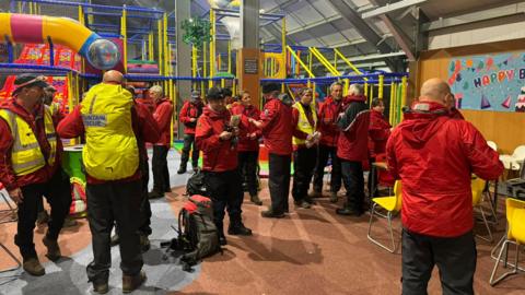 Rescuers dressed in red and yellow clothing seen gathering at a child's play centre with soft play activities in the background. There are around 14 people in the room.