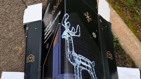 The box of the light up Christmas decoration. There is a a silver reindeer with lights around it and gold snowflakes around it. 