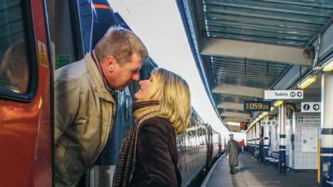 A man on a train and a woman on the platform lean in to each other as if to kiss.