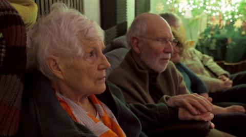 An elderly lady with short grey hair and an orange scarf. She is sitting on a sofa next to an elderly man who is wearing glasses. The man is resting his hand on top of the lady's hand. They are looking into the distance. There are Christmas lights in the background. 