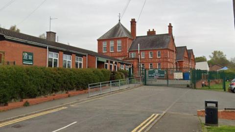 A red brick Victorian building with a green gate in front of it. A long row of hedges run along the side of the building. 