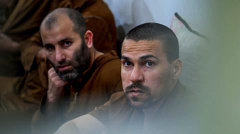 Two inmates sit on the ground against a wall both dressed in brown clothes