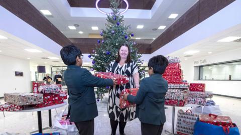 Two boys dressed in green blazers in a room full of wrapped Christmas presents and a Christmas tree. They are each handing a gift to a woman dressed in a black and white dress, who is stood in the middle of them