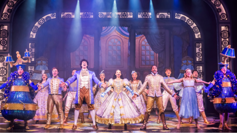 The cast of Cinderella, dressed in big dresses and fancy waist coats, stand on a stage singing